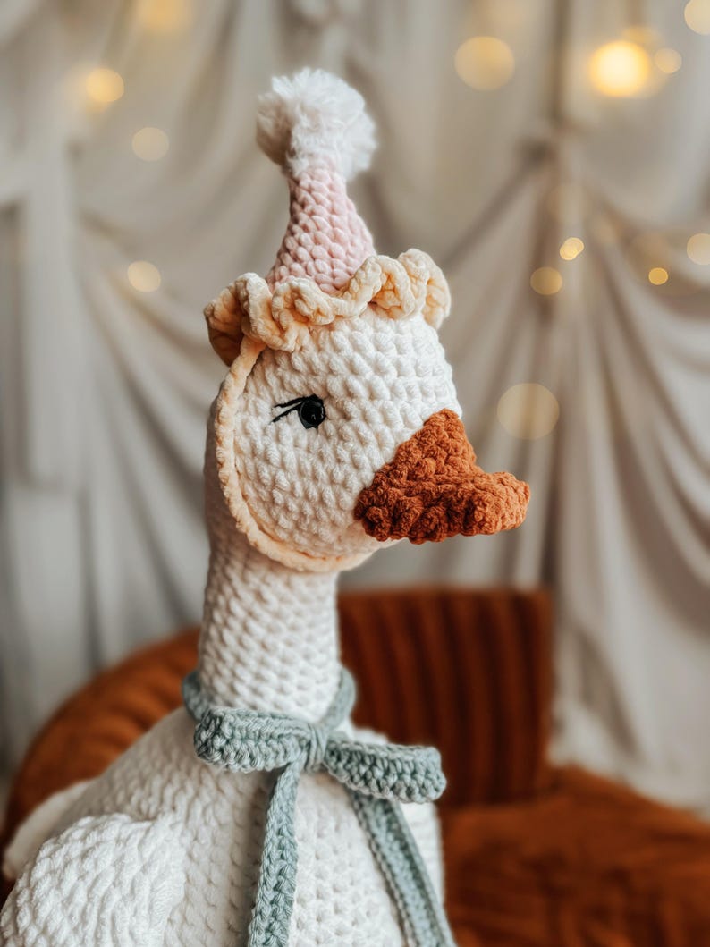 May include: A handmade, white crochet goose wearing a party hat with a pink and white gradient and a light green bow tie. The goose has an orange beak and black eyes. The plush toy is designed for children.