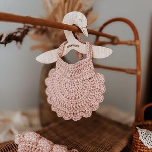 May include: A light pink, crocheted baby dress hangs on a wooden swan-shaped hanger. The hanger is suspended from a wooden rod. The dress has a scalloped edge. The word "BABYBOOPS" is engraved on the hanger.