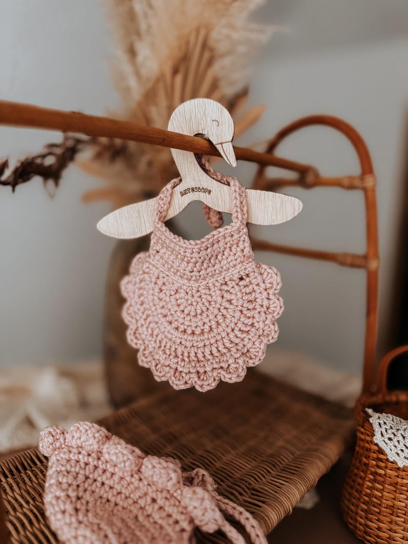 May include: A light pink, crocheted baby dress hangs on a wooden swan-shaped hanger. The hanger is suspended from a wooden rod. The dress has a scalloped edge. The word "BABYBOOPS" is engraved on the hanger.