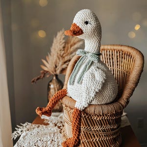 May include: A handmade, white crochet goose doll with an orange beak and feet, and a green bow tie. The goose is seated in a wicker chair. The background includes a vase with dried plants and soft lighting.