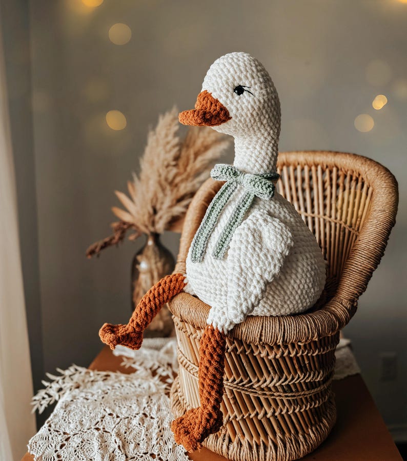 May include: A handmade, white crochet goose doll with an orange beak and feet, and a green bow tie. The goose is seated in a wicker chair. The background includes a vase with dried plants and soft lighting.