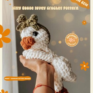 Pu&ograve; includere: Un giocattolo di oca all'uncinetto con un fiocco marrone, un corpo bianco e un becco arancione. L'oca &egrave; tenuta da una mano con un braccialetto rosso. L'immagine include il testo "Silly Goose Lovey Crochet Pattern" e "Low Sew".