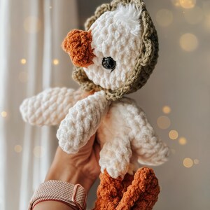 Pu&ograve; includere: Un giocattolo di peluche a forma di oca lavorato a uncinetto con un corpo bianco, gambe marroni e una testa con cappuccio verde.