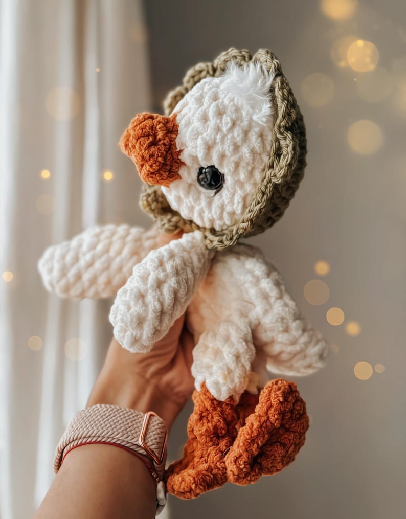Pu&ograve; includere: Un giocattolo di peluche a forma di oca lavorato a uncinetto con un corpo bianco, gambe marroni e una testa con cappuccio verde.