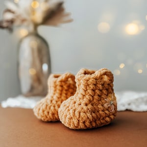 May include: A pair of handmade, light brown crochet baby booties. The booties are made with a thick yarn, giving them a textured appearance. The background is blurred, with a vase of dried flowers and soft lighting.