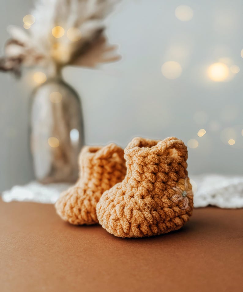 May include: A pair of handmade, light brown crochet baby booties. The booties are made with a thick yarn, giving them a textured appearance. The background is blurred, with a vase of dried flowers and soft lighting.