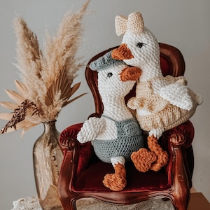 May include: Two handmade crochet geese, one wearing a blue cap and overalls, the other a bow and dress, sit on a red velvet chair. The geese have orange beaks and feet. A vase with dried plants is in the background.