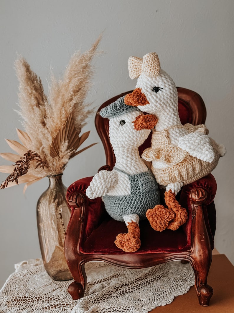 May include: Two handmade crochet geese, one wearing a blue cap and overalls, the other a bow and dress, sit on a red velvet chair. The geese have orange beaks and feet. A vase with dried plants is in the background.