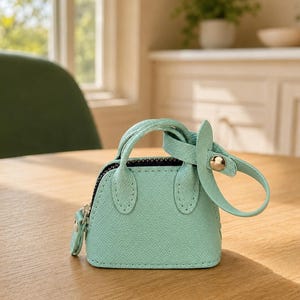 May include: A miniature mint green handbag featuring a textured faux leather finish, two small top handles, a top zipper closure, and a detachable strap with a gold-toned metal snap button fastener attached to one of the handles.