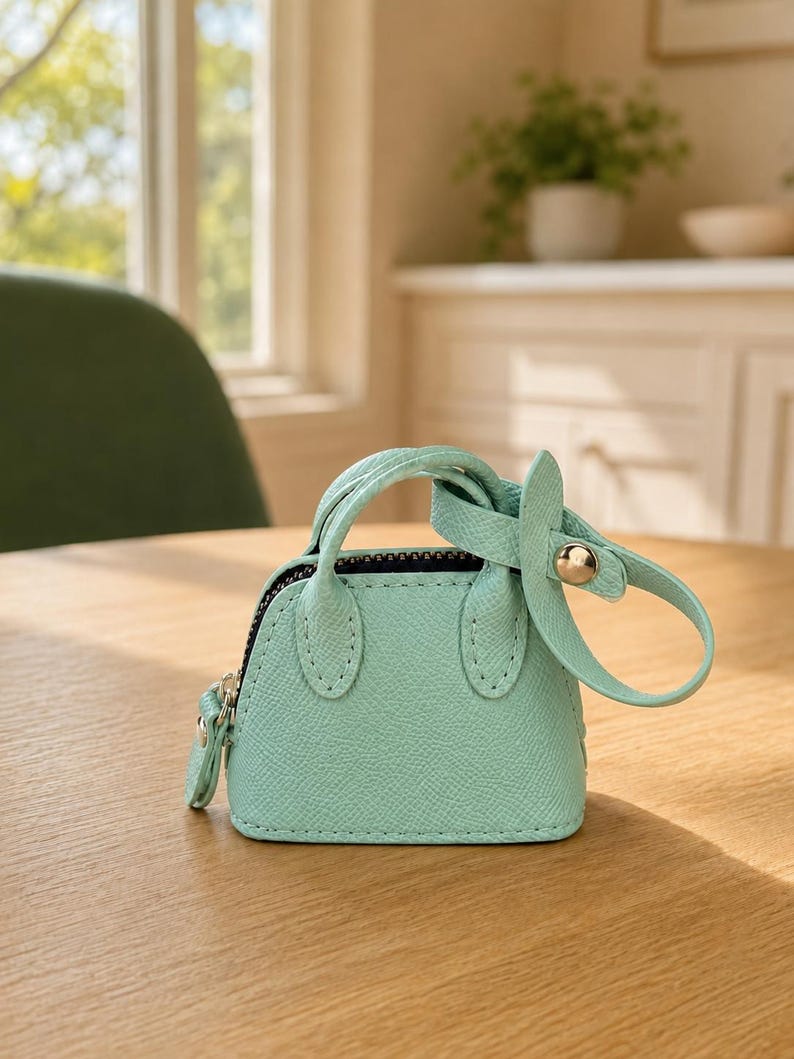 May include: A miniature mint green handbag featuring a textured faux leather finish, two small top handles, a top zipper closure, and a detachable strap with a gold-toned metal snap button fastener attached to one of the handles.