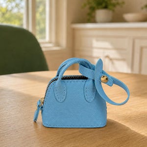 May include: A miniature light blue handbag charm with a textured surface, featuring two small handles, a top zipper closure, and a detachable strap secured with a gold-toned metal snap button. The accessory has a structured, dome-shaped silhouette.