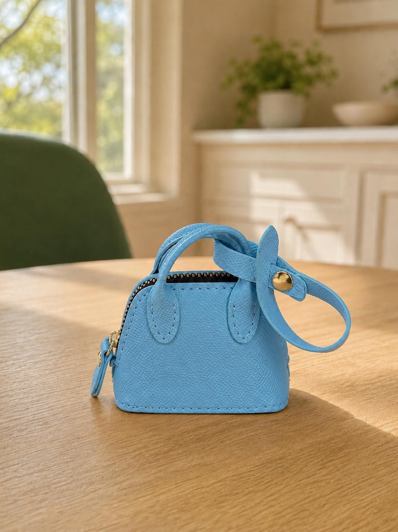 May include: A miniature light blue handbag charm with a textured surface, featuring two small handles, a top zipper closure, and a detachable strap secured with a gold-toned metal snap button. The accessory has a structured, dome-shaped silhouette.
