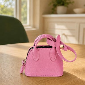 May include: A miniature bright pink handbag featuring a textured surface, two small top handles, a black zipper closure, and a detachable strap with a gold-toned metal snap button fastener attached to one of the handles.