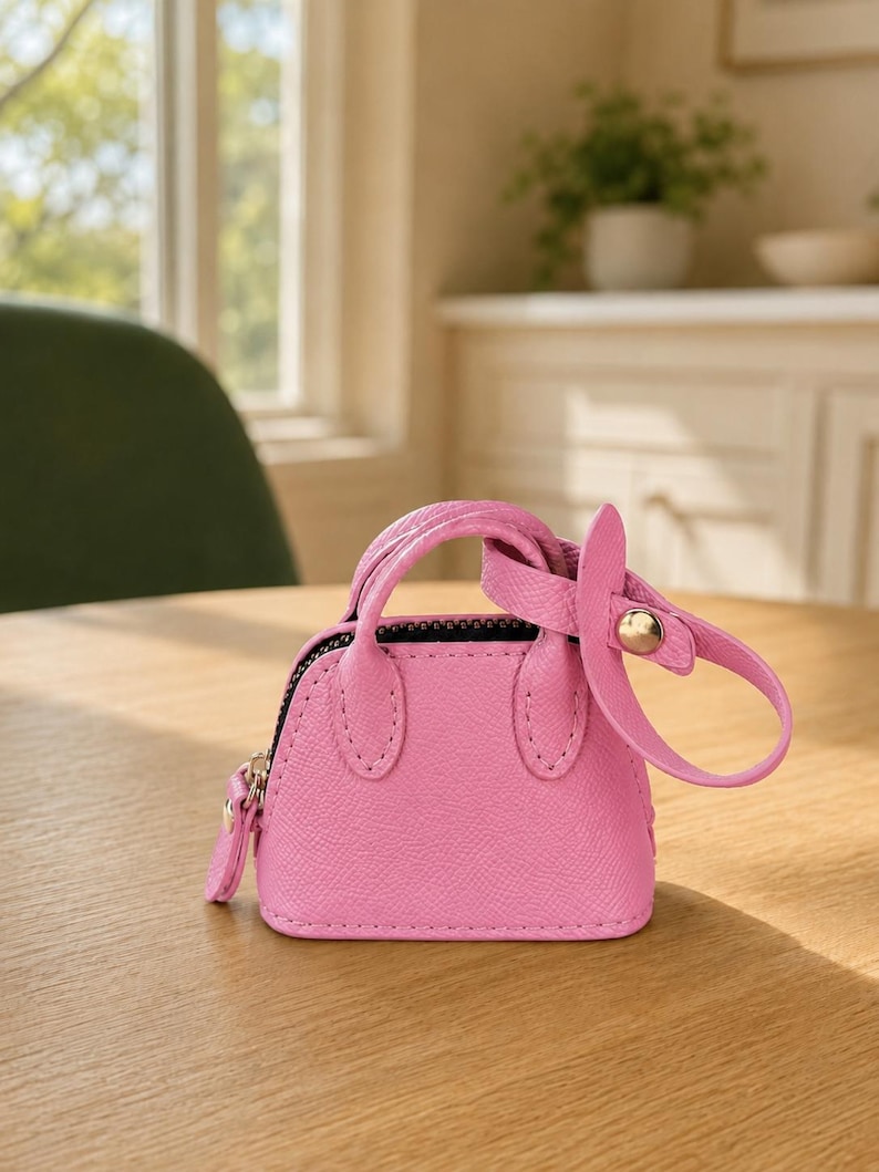 May include: A miniature bright pink handbag featuring a textured surface, two small top handles, a black zipper closure, and a detachable strap with a gold-toned metal snap button fastener attached to one of the handles.