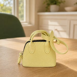 May include: A miniature pale yellow handbag charm with a textured surface, two small handles, a black zipper closure, and a matching yellow strap with a gold-toned metal snap button fastener attached to the side of the bag.