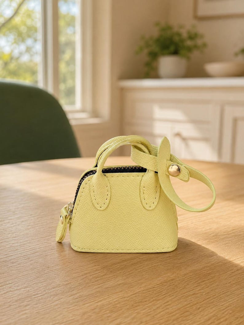May include: A miniature pale yellow handbag charm with a textured surface, two small handles, a black zipper closure, and a matching yellow strap with a gold-toned metal snap button fastener attached to the side of the bag.