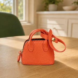 May include: A miniature orange handbag with a textured surface, featuring two short top handles, a black zipper closure, and a detachable strap with a gold-toned metal snap button fastener attached to one of the handles.