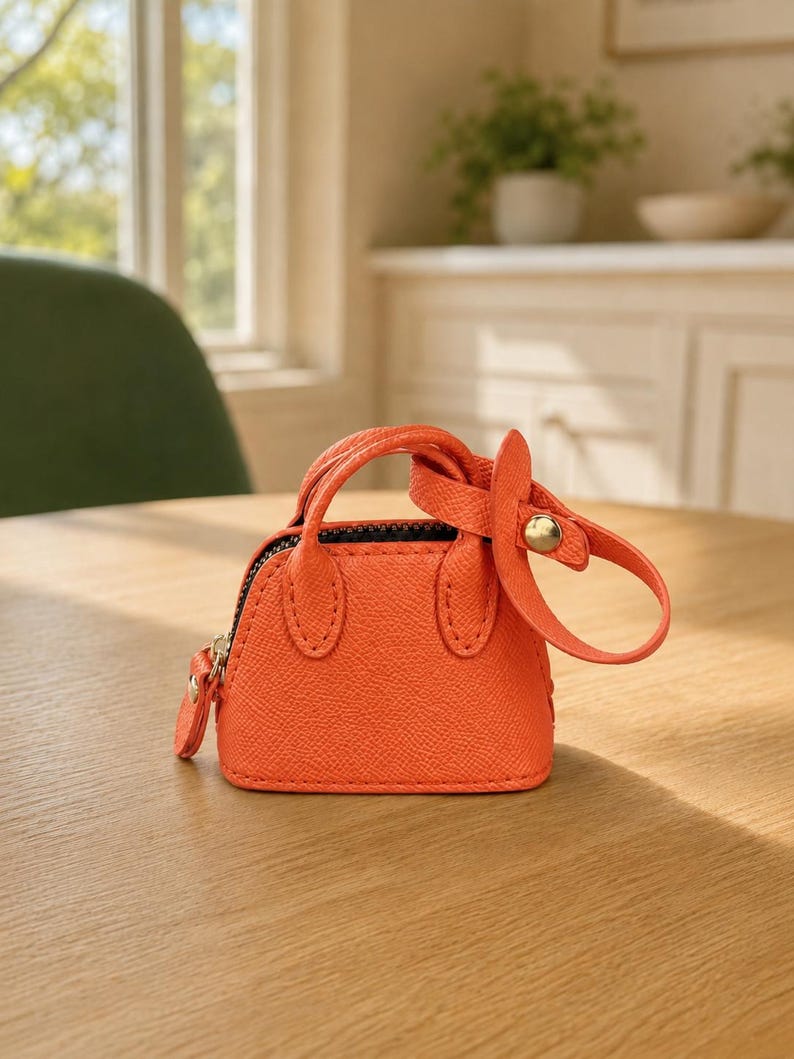 May include: A miniature orange handbag with a textured surface, featuring two short top handles, a black zipper closure, and a detachable strap with a gold-toned metal snap button fastener attached to one of the handles.