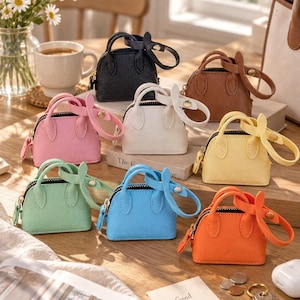 May include: A collection of eight miniature handbags in black, brown, pink, white, yellow, mint green, blue, and orange. Each small bag features a top handle, a zipper closure, and a matching wrist strap attached with a gold-toned metal stud.