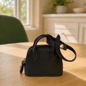 May include: A miniature black handbag with a textured surface, featuring two small top handles, a top zipper closure, and a detachable strap with a gold-toned metal snap button. The bag has a structured, dome-shaped silhouette.