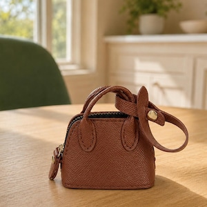 May include: A miniature brown handbag with a textured surface, featuring two small top handles, a zipper closure along the top, and a detachable strap with a gold-toned metal snap button fastener attached to one of the handles.