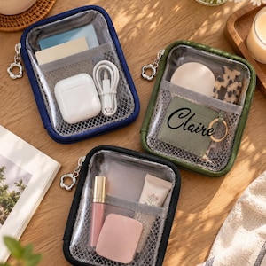 May include: Three rectangular zippered pouches with clear front panels and mesh interior pockets. One pouch is navy blue, one is olive green with the name Claire printed on a small pocket, and one is black. Each features a silver flower-shaped zipper pull.