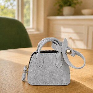 May include: A miniature light blue handbag charm with a textured surface, two small handles, a black zipper closure, and a matching light blue strap attached with a gold-toned metal stud. The accessory features a structured dome shape.