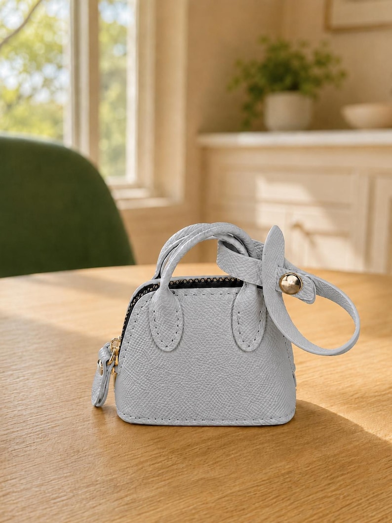 May include: A miniature light blue handbag charm with a textured surface, two small handles, a black zipper closure, and a matching light blue strap attached with a gold-toned metal stud. The accessory features a structured dome shape.