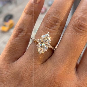 Op de afbeelding: Een verlovingsring met een marquise-geslepen diamant. De heldere diamant is gezet in een gouden band met kleinere accentstenen. De ring zit aan een hand met verzorgde nagels.