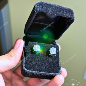 A hand holding a black jewelry box containing two diamond earrings.