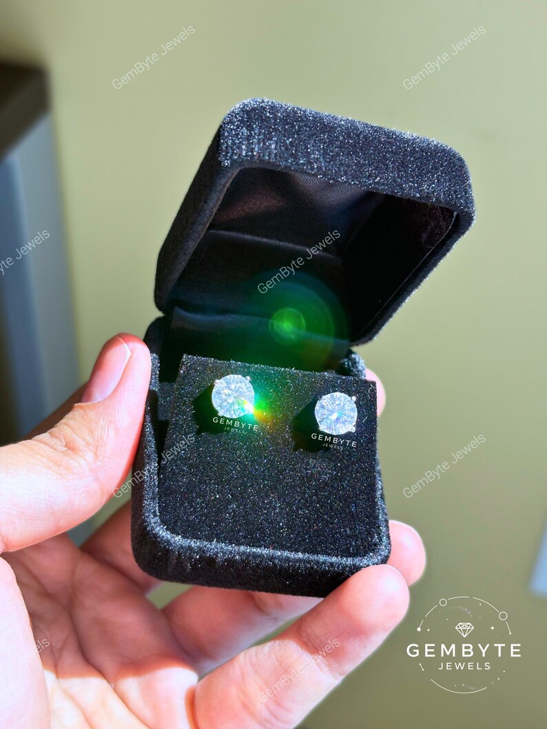 A hand holding a black jewelry box containing two diamond earrings.