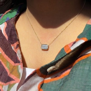 May include: A gold necklace featuring a rectangular, emerald-cut diamond pendant. The pendant is set in a gold bezel and hangs from a delicate gold chain. The necklace is displayed against a colorful patterned shirt.