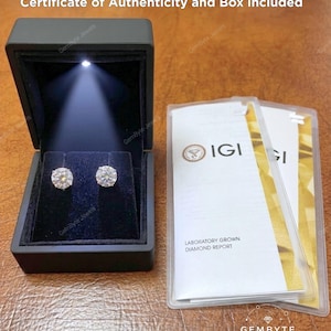 a pair of diamond earrings in a black jewelry box, accompanied by a certificate of authenticity and a box containing a laboratory grown diamond report.