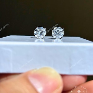 A pair of diamond earrings being held in a hand, with the earrings resting on a white surface.
