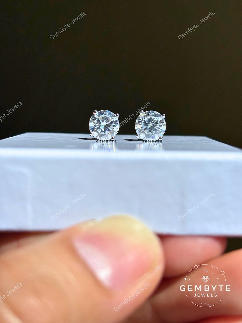 A pair of diamond earrings being held in a hand, with the earrings resting on a white surface.