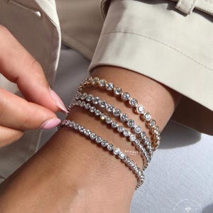 Könnte beinhalten: Vier Tennisarmbänder mit runden, in Zargenfassung gefassten klaren Steinen in einer durchgehenden Reihe. Die Armbänder sind in verschiedenen Metalltönen wie Gold und Silber gefertigt und tragen den Schriftzug GEMBYTE JEWELS.
