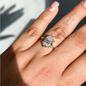 May include: A gold ring featuring a large, oval-cut, faceted clear gemstone held by four prongs on a simple, polished gold band. The ring is positioned on a finger, and the text GEMBYTE JEWELS is visible near the setting and in the corner.