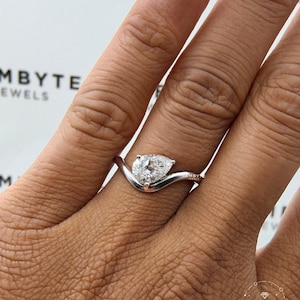 May include: A silver-toned ring featuring a pear-shaped clear gemstone set on a curved, wave-like band. The metal band has a smooth, polished finish, and the setting holds the stone securely at the center of the V-shaped curve of the band.