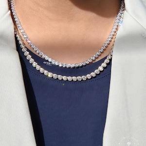 May include: Two layered tennis necklaces featuring round, bezel-set clear stones. The top necklace is silver-toned, and the bottom necklace is gold-toned. Both necklaces feature the text GEMBYTE JEWELS printed in small white lettering.