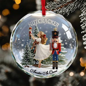 May include: A clear glass Christmas bauble with a red ribbon. Inside, a scene from The Nutcracker ballet features a ballerina, a nutcracker soldier, and snowy pine trees. The bauble is inscribed with "NUTCRACKER" and "Amelia As Clara".