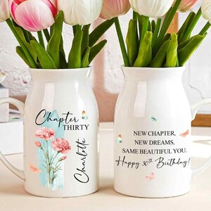 May include: Two white ceramic vases with handles, each holding a bouquet of pink and white tulips. One vase reads "Chapter Thirty Charlotte" and the other reads "Happy 30th Birthday!" with a quote. The vases have floral and butterfly designs.