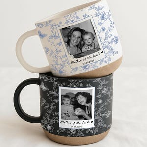May include: Two ceramic mugs stacked on top of each other. The top mug is white with blue floral patterns and a photo that says "Mother of the Bride" and the date 03.21.2026. The bottom mug is black with white floral patterns and a photo that says "Mother of the Bride" and the date 04.15.2026.