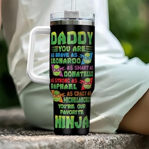 May include: A black stainless steel tumbler with a white handle and lid. The tumbler features a graphic of the Teenage Mutant Ninja Turtles characters and the text "Daddy You Are As Brave As Leonardo As Smart As Donatello As Strong As Raphael As Crazy As Michelangelo You're Our Favorite Ninja".