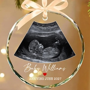 May include: A circular glass ornament featuring a black and white ultrasound image of a fetus. The text reads Baby Williams with a small red heart icon below, followed by the text ARRIVING JUNE 2027. A light beige satin ribbon is tied at the top.