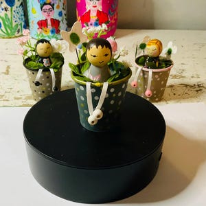 Può includere: Tre piccoli secchi decorativi in metallo a pois, ciascuno contenente una figura di bambola di legno e fiori artificiali. I secchi sono in tonalità di verde e marrone, con dettagli di nastro bianco. Le bambole hanno volti dipinti.