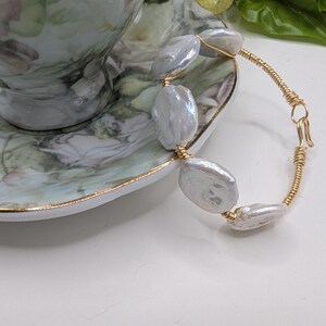 Peut inclure: Un bracelet délicat orné de grandes perles blanches nacrées irrégulières, enfilées sur un fil de couleur or. Le bracelet est présenté près d'une tasse et d'une soucoupe à motifs floraux, suggérant un thème d'élégance et de raffinement.