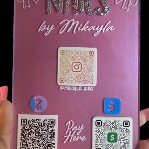 May include: A pink acrylic sign with the word "NAILS" in rhinestone letters and "by Mikayla" in white script. It features QR codes, social media handles, and payment options.