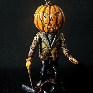 May include: A Halloween-themed figurine of a person with a carved pumpkin head. The pumpkin is orange with a scary face and the person is wearing a brown jacket and black pants. The figurine is standing on a wooden base.