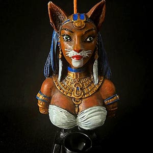 May include: A ceramic bust of a cat-like woman with blue and gold jewelry, wearing a white top and a blue headdress. The bust is on a black base.
