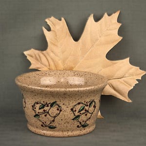 May include: A speckled beige ceramic bowl with heart-shaped vine designs in brown and green. A large, tan leaf rests behind the bowl, creating a rustic, autumnal aesthetic. The bowl is likely used for serving or display.